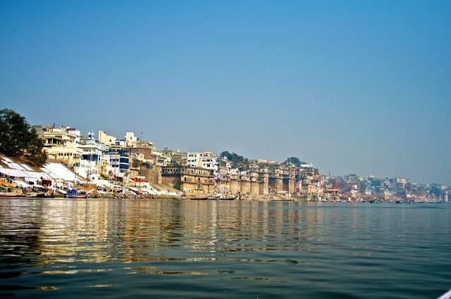 Un denso paisaje urbano de edificios tradicionales y ghats a lo largo de un río ancho, visto desde el agua bajo un cielo azul claro.