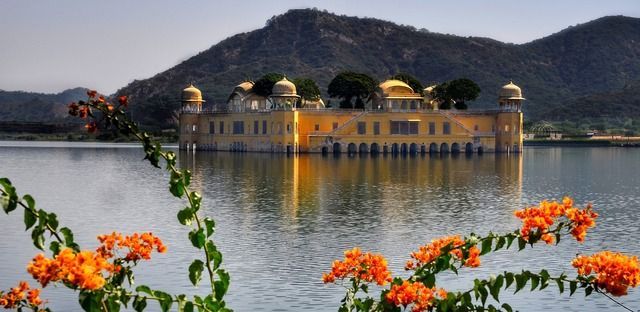 Un palacio amarillo ornamentado se asienta en un lago con una montaña detrás, enmarcado por flores naranjas.