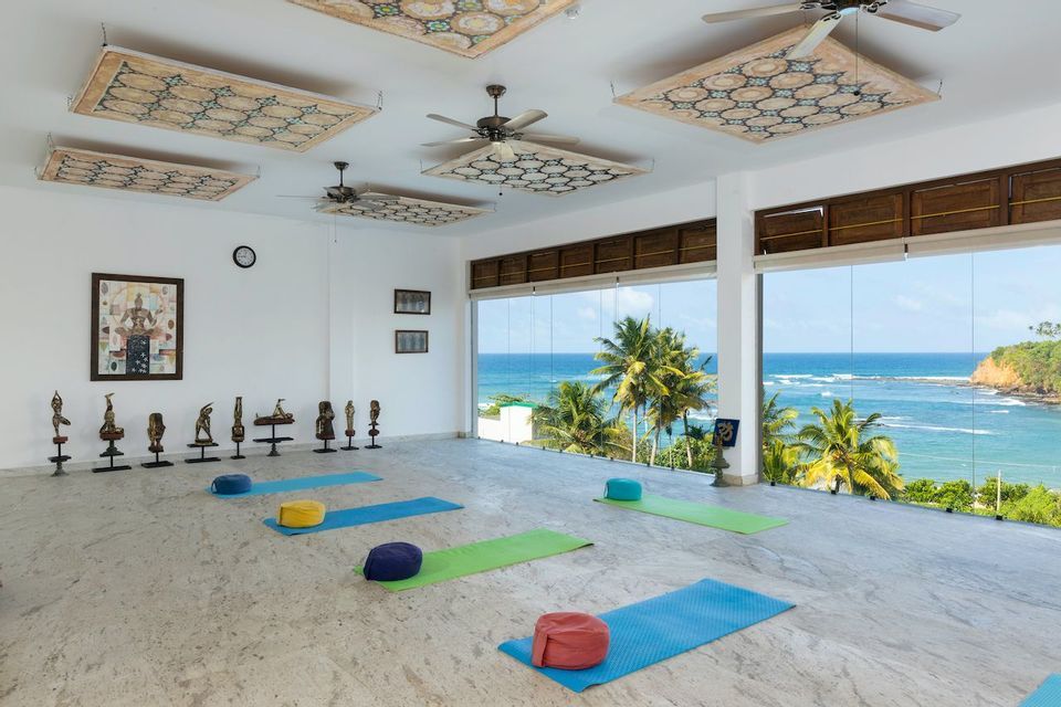 Uno studio yoga luminoso e vuoto con tappetini e cuscini su un pavimento di marmo, con ampie finestre che mostrano un oceano tropicale e palme.