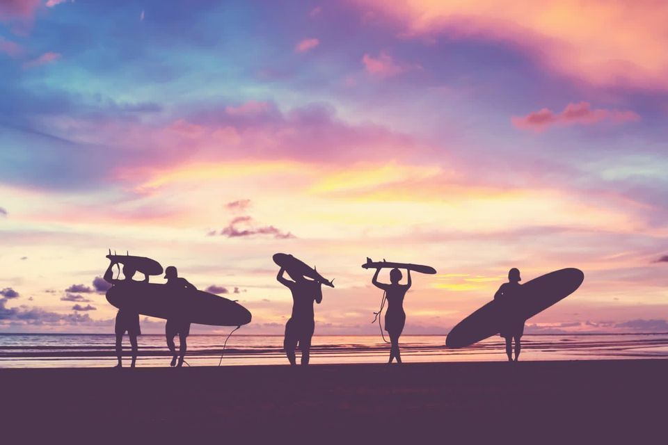 Sagome di un gruppo WeRoad con tavole da surf su una spiaggia al tramonto, sotto un cielo colorato.