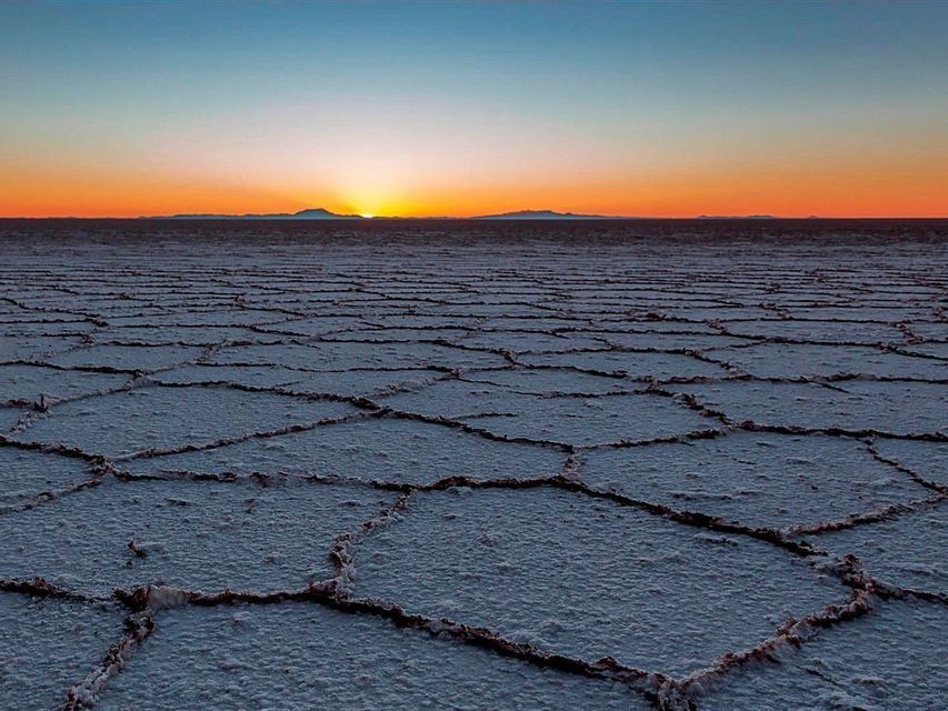 El sol se pone sobre un vasto salar agrietado, creando un resplandor naranja en el horizonte con montañas distantes.