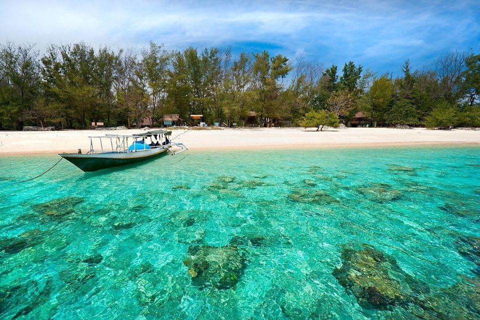 Un bateau flotte dans une eau turquoise peu profonde et transparente, révélant des rochers sur le fond marin, près d'une plage de sable bordée d'arbres.