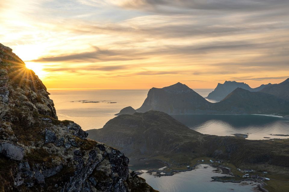 Un raggio di sole spunta da dietro una montagna al tramonto, illuminando un paesaggio costiero con fiordi e vette distanti sotto un cielo nuvoloso.