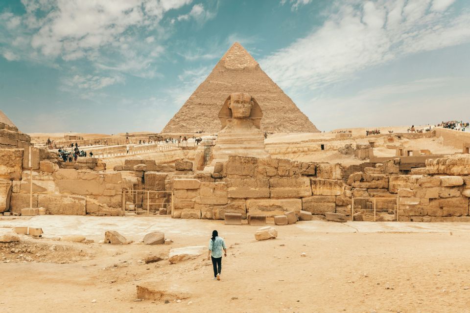 Eine Person geht durch sandige Ruinen auf die Sphinx zu, im Hintergrund ist eine große Pyramide unter einem bewölkten blauen Himmel sichtbar.