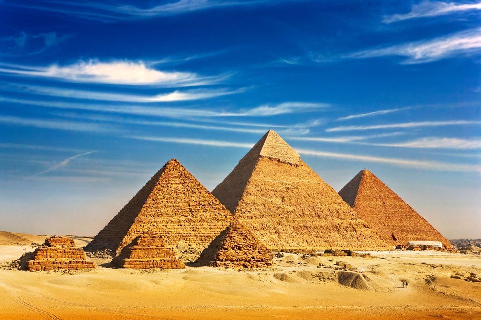 Diverse grandi piramidi di pietra e rovine più piccole si ergono in un deserto sabbioso sotto un cielo blu vibrante con nuvole leggere.