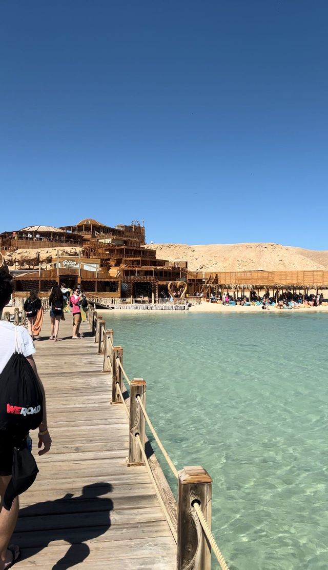 Un viaggio di gruppo WeRoad cammina lungo un molo di legno su acqua cristallina e turchese verso una spiaggia sabbiosa e un grande edificio del resort.