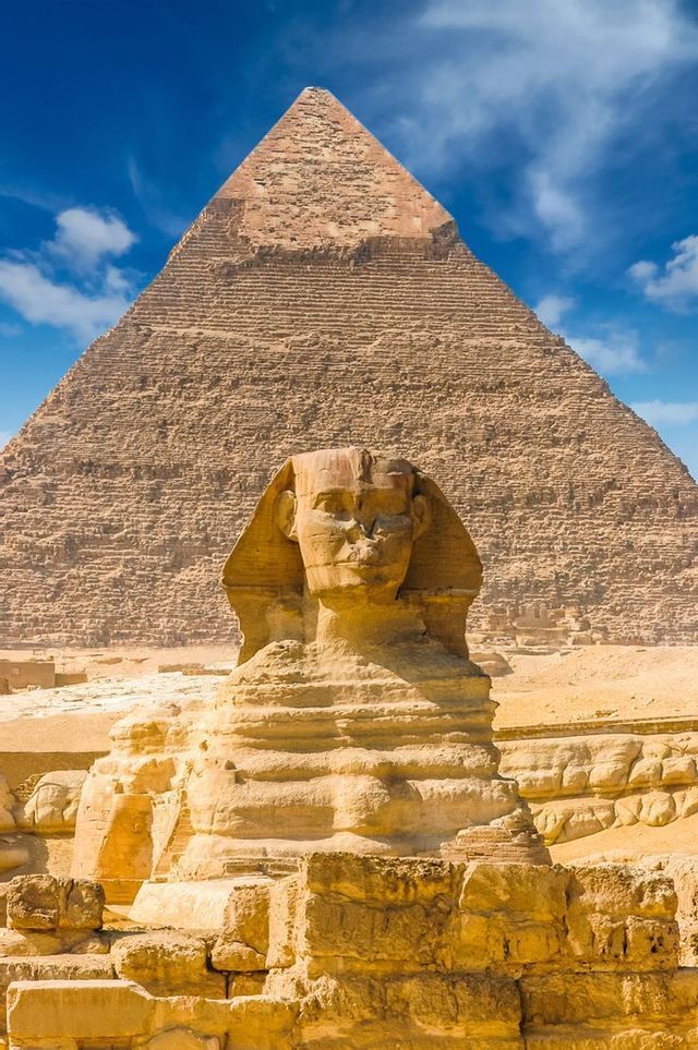 La Grande Sfinge si erge davanti a una grande piramide nel deserto sotto un cielo azzurro con nuvole.
