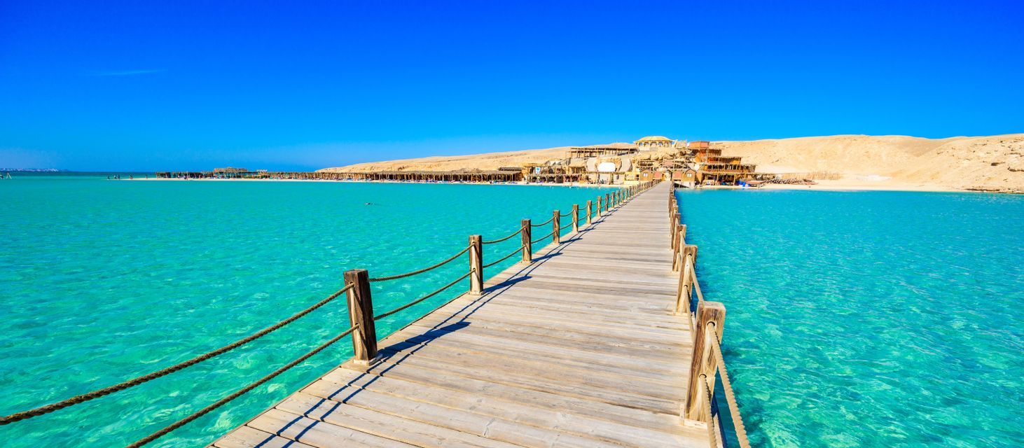 Un molo di legno con ringhiere in corda si estende su acque limpide color turchese fino a una spiaggia sabbiosa con edifici ai piedi di una duna.