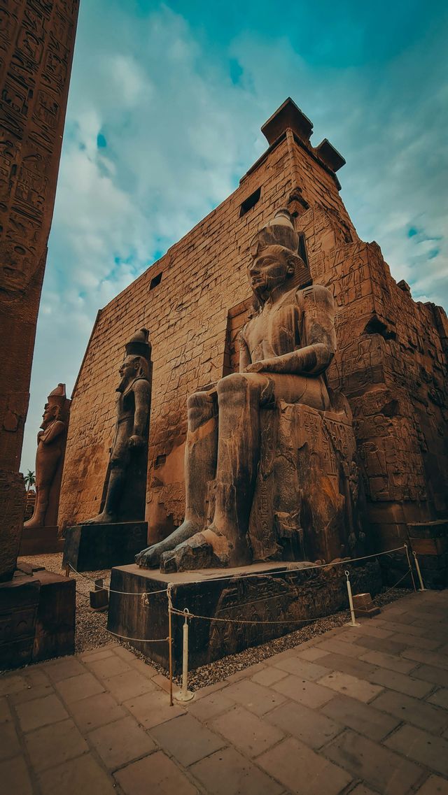 Un'inquadratura dal basso di colossali statue egizie antiche, sedute e in piedi, davanti a una parete del tempio sotto un cielo nuvoloso.