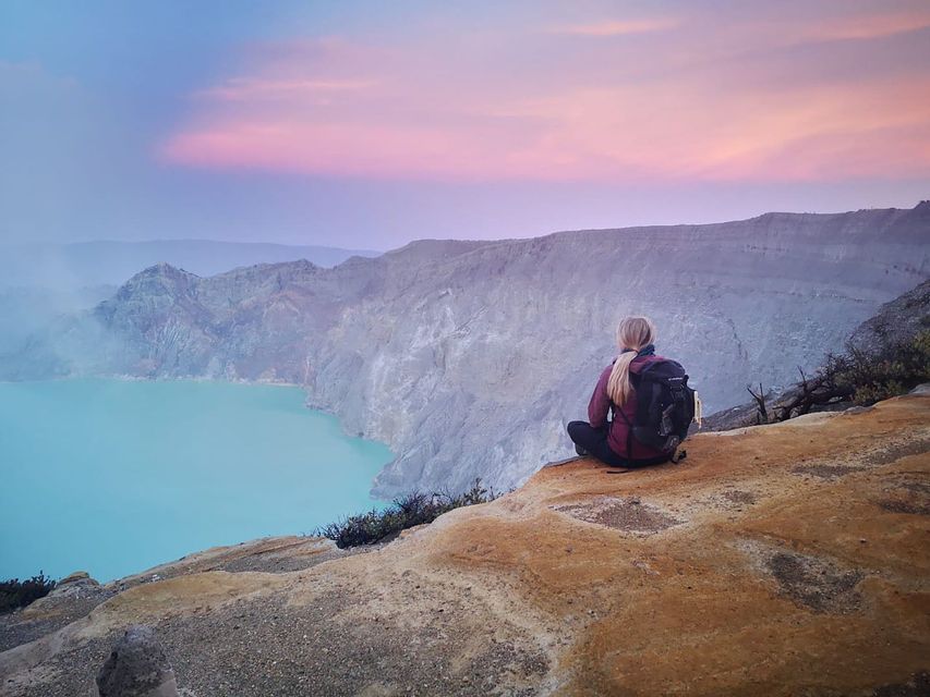 Un randonneur est assis au bord d'une falaise rocheuse, surplombant un lac de cratère turquoise sous un ciel rose et bleu.