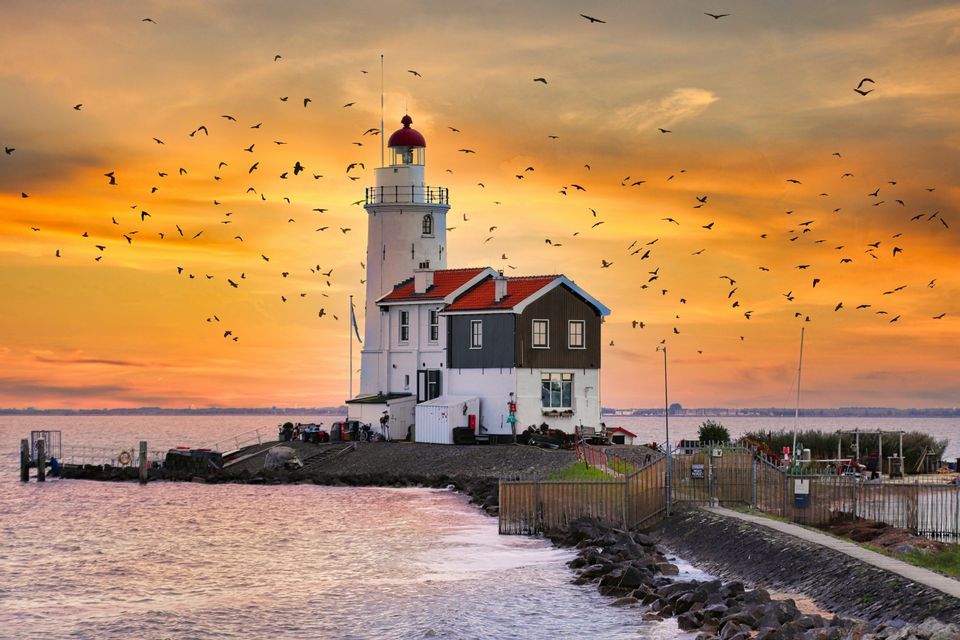 Un faro blanco con techo rojo se alza en una costa rocosa, mientras una bandada de pájaros vuela por el cielo naranja al atardecer.
