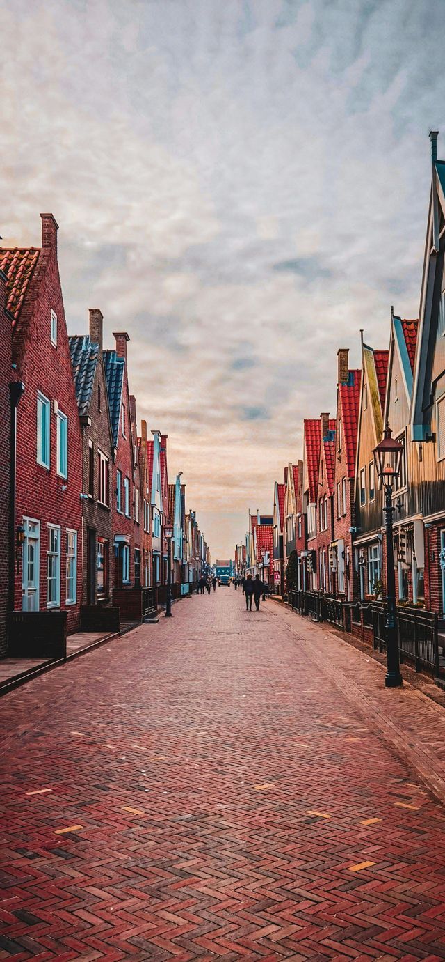 Una calle larga y recta pavimentada con ladrillos rojos, flanqueada por casas altas y estrechas con tejados a dos aguas bajo un cielo nublado.