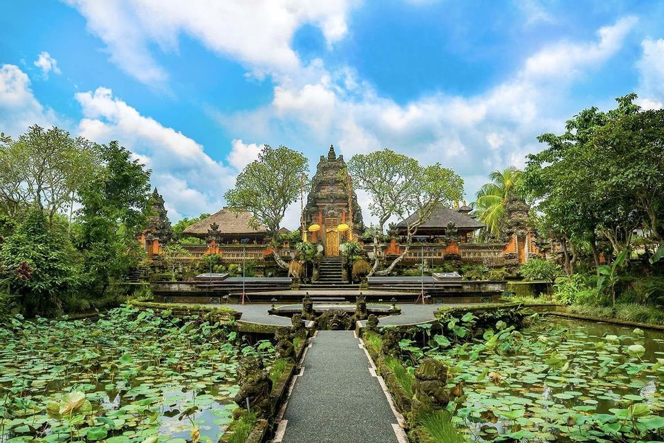 Un sentiero in pietra conduce su uno stagno coperto di ninfee fino a un tempio ornato circondato da alberi sotto un cielo blu e bianco.