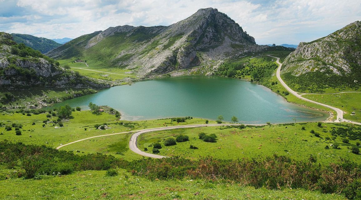 Un lago turquesa se asienta en un valle rodeado de montañas verdes y rocosas, con una carretera sinuosa que bordea su orilla bajo un cielo parcialmente nublado.