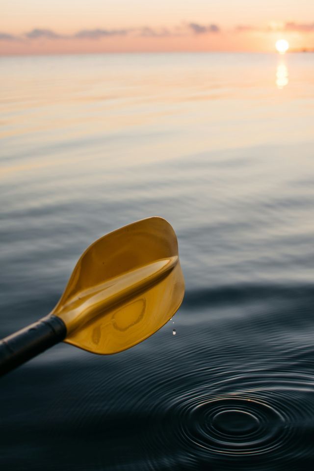Un remo amarillo gotea agua sobre una superficie tranquila, creando ondas con el atardecer de fondo.