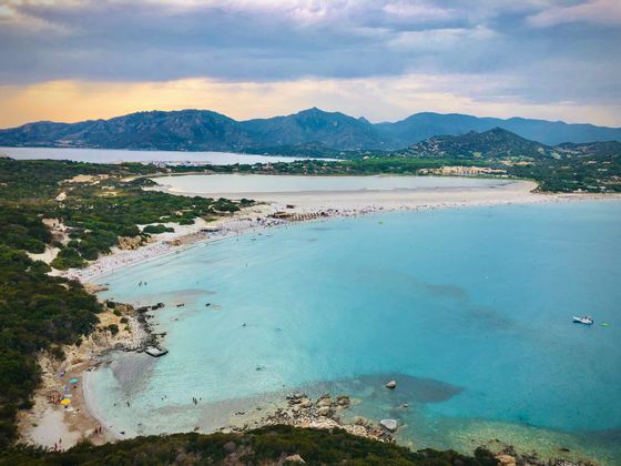 Vista dall'alto di una lunga spiaggia sabbiosa che separa una baia turchese da una laguna calma, con montagne in lontananza.