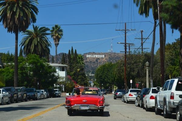 Une décapotable rouge vintage roule le long d'une rue bordée de palmiers, avec vue sur l'enseigne Hollywood sur une colline lointaine.