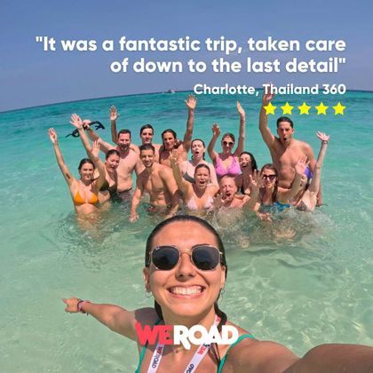 A woman takes a selfie with a WeRoad group trip, with everyone smiling and waving from the turquoise sea.