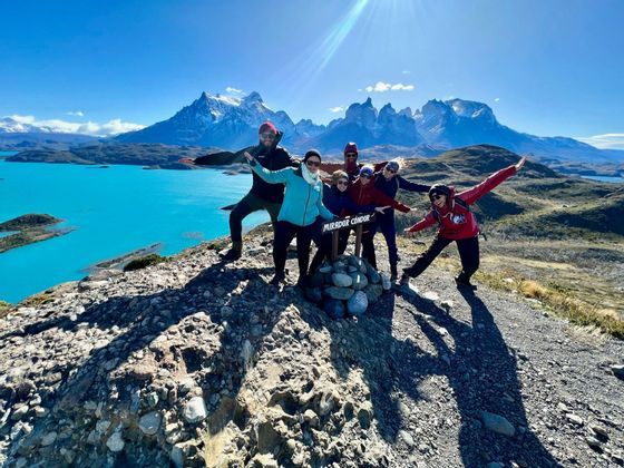 A WeRoad group trip poses on a rocky viewpoint overlooking a turquoise lake and a snowy mountain range under a clear sky.
