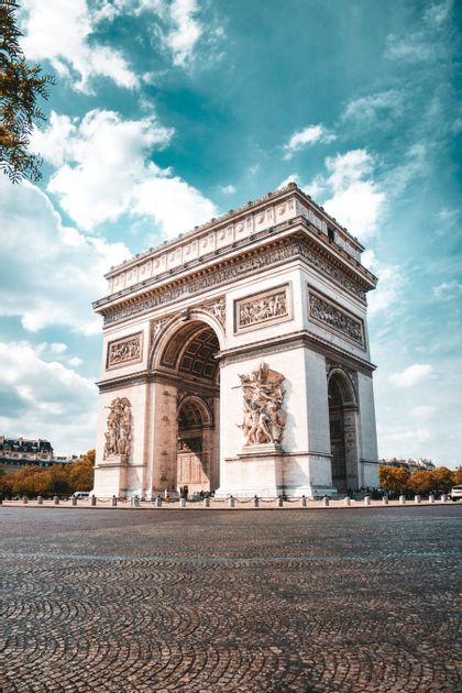 Un grande e ornato arco trionfale si erge su un'ampia piazza lastricata sotto un cielo azzurro con nuvole sparse.