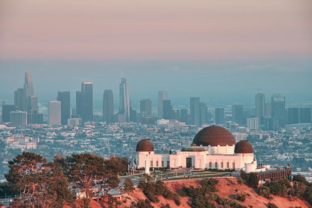 An observatory with large domes sits on a hillside, overlooking a sprawling city skyline with skyscrapers at sunset.