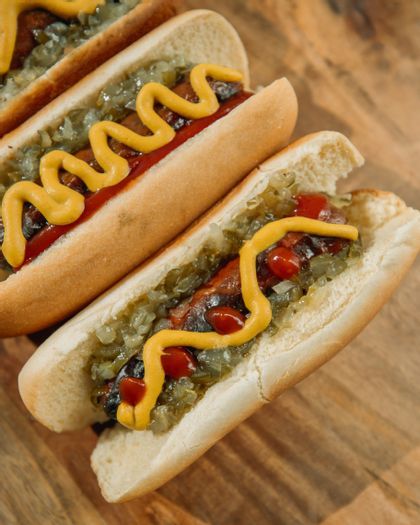 Primo piano di due hot dog nei panini, conditi con relish verde, ketchup e una striscia a zig-zag di senape gialla, su un tagliere di legno.