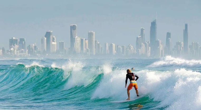 Un surfista cavalca una grande onda turchese nell'oceano con lo skyline di una città lontana e sfocata sullo sfondo.
