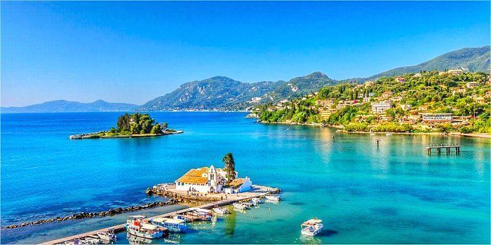Un monastère blanc sur un petit îlot, relié à une jetée, situé dans une baie aux eaux turquoise éclatantes et un littoral verdoyant et vallonné.