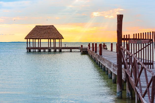 Un muelle de madera conduce a un pabellón con techo de paja sobre aguas tranquilas bajo un cielo al atardecer con rayos de sol.