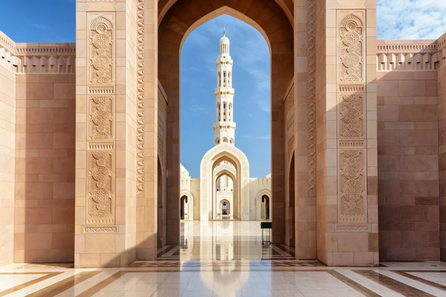 L'alto minareto di una moschea è incorniciato da un grande arco in pietra, con il suo riflesso visibile sul pavimento lucido del cortile.