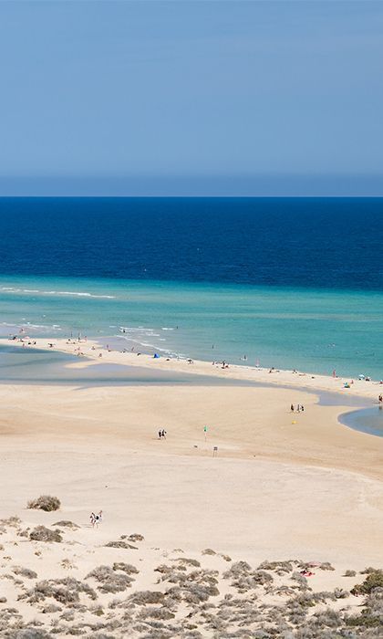 An elevated view of a sandy spit beach with people, separating turquoise water from the deep blue sea.