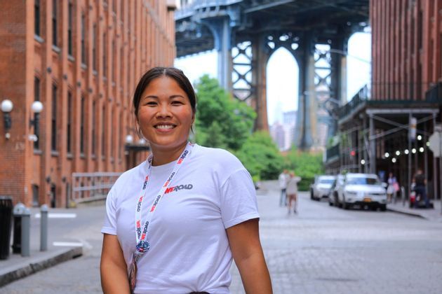 A smiling woman in a WeRoad t-shirt stands on a cobblestone street with a large bridge and brick buildings in the background.