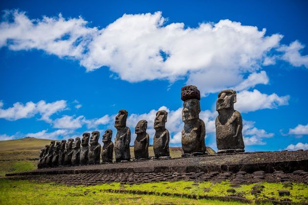 Una fila di imponenti statue Moai si erge su una piattaforma di pietra, su un prato verde sotto un cielo azzurro luminoso con nuvole.