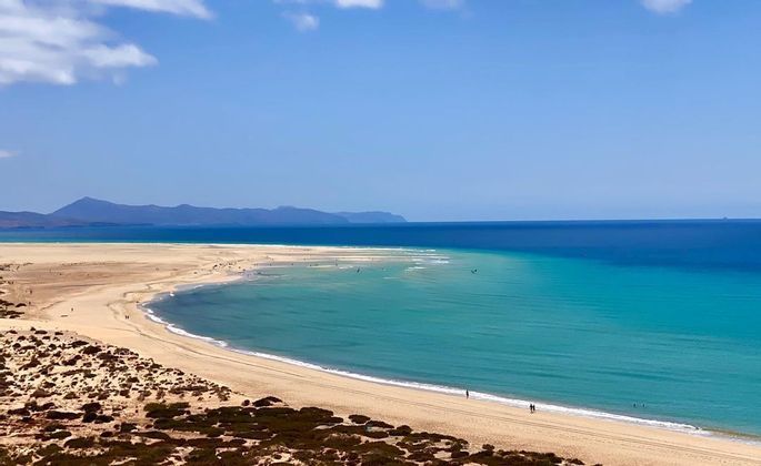 Una vista panoramica di una lunga spiaggia sabbiosa e curva che incontra acque turchesi e blu intenso, con montagne in lontananza sotto un cielo azzurro.