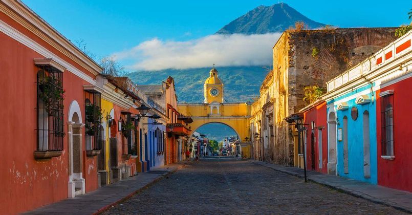 Un arco de torre de reloj amarilla se alza sobre una calle adoquinada, flanqueada por edificios coloridos y con una gran montaña al fondo.