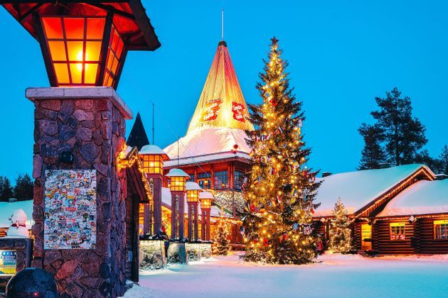 Un grande albero di Natale decorato con luci si erge in un villaggio innevato con edifici illuminati al crepuscolo.