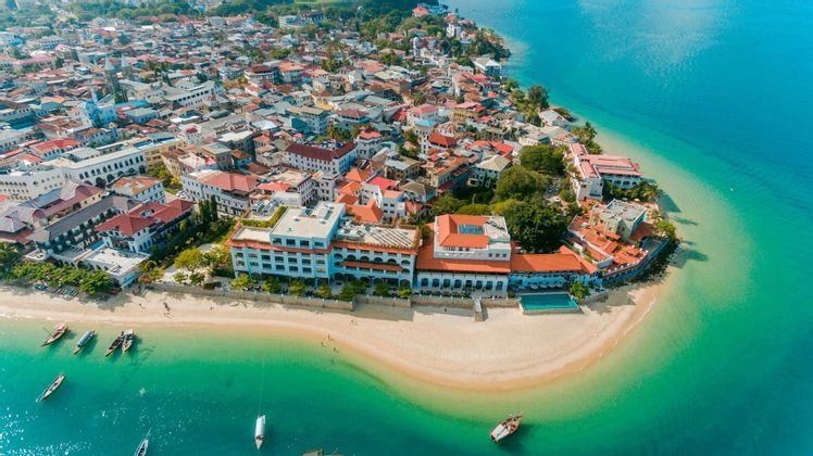 Luftaufnahme einer Küstenstadt auf einer Halbinsel mit einem Sandstrand, umgeben von klarem türkisfarbenem Wasser mit Booten.