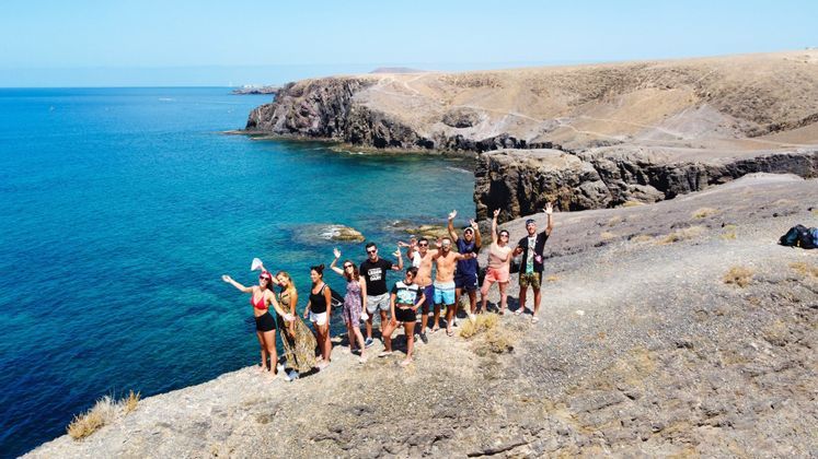 Un gruppo WeRoad in posa saluta da una scogliera rocciosa che domina un mare turchese e una costa arida e collinare.