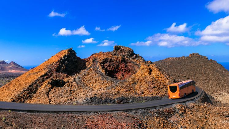 Un autobus turistico arancione percorre una strada tortuosa attraverso un vasto paesaggio vulcanico con crateri sotto un cielo azzurro e limpido.