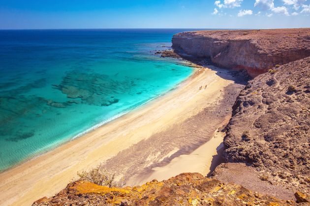 Una vista dall'alto di un viaggio di gruppo WeRoad che cammina su una spiaggia sabbiosa accanto ad acqua turchese e grandi scogliere bruno-rossastre.