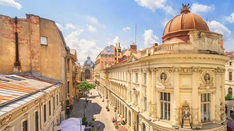 Vista en picada de una calle de ciudad soleada, bordeada por edificios clásicos ornamentados y una gran cúpula naranja bajo un cielo azul.