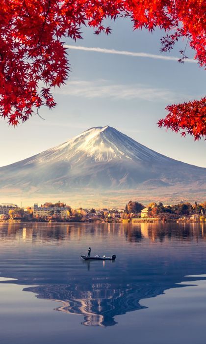 Une montagne enneigée se reflète sur un lac calme où une personne se tient dans une petite embarcation, encadrée par des feuilles d'automne rouges.