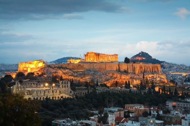 Un'antica cittadella, splendidamente illuminata da luci dorate, si erge su una collina rocciosa che domina un paesaggio urbano al tramonto.