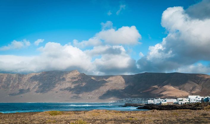 A coastal town with white buildings sits by the sea at the foot of large mountains under a partly cloudy sky.