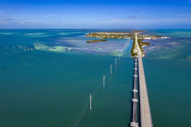 Luftaufnahme einer langen Brücke, die über türkisfarbenes Meerwasser zu einer kleinen Insel führt, mit Strommasten daneben.