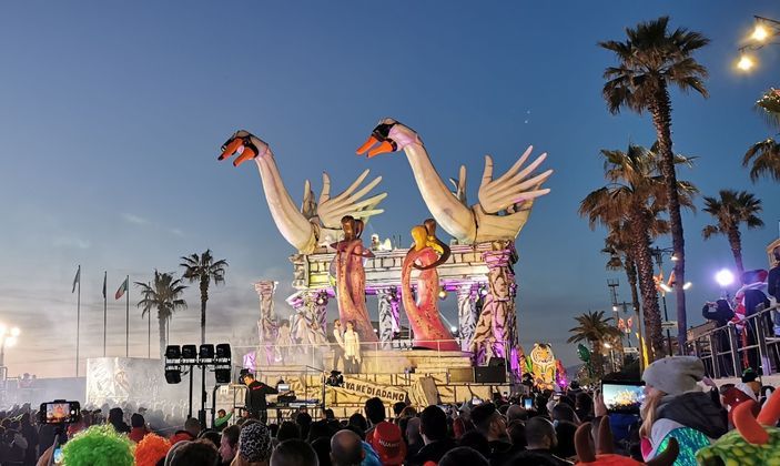 A large, ornate carnival float with swan sculptures moves through a crowd of people during a parade at dusk.