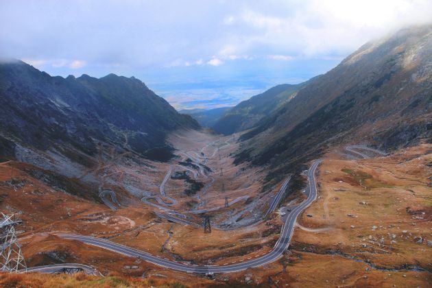 Vista aérea de una carretera sinuosa con muchas curvas cerradas descendiendo por un gran valle de montaña bajo un cielo nublado.