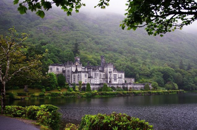 Un grand château de pierre se dresse sur les rives d'un lac, avec une colline escarpée et densément boisée s'élevant dans la brume en arrière-plan.