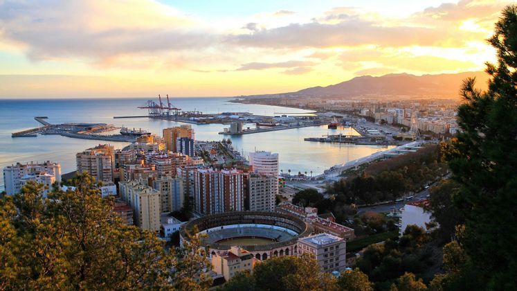 Vista panoramica di una città costiera con arena, porto commerciale e montagne sullo sfondo al tramonto.