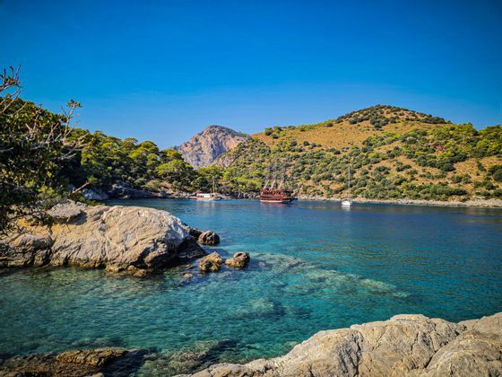 Una grande nave di legno e altre imbarcazioni ancorate in una baia tranquilla con acqua cristallina turchese e colline rocciose e verdi sotto un cielo azzurro brillante.